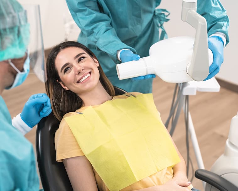 Young woman in yellow bib with white teeth smiling in dentist's chair with two dentists over her preparing to take x-rays of her teeth