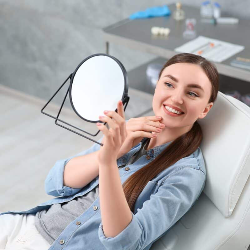 Woman smiling, holding mirror in dental office after orthodontics treatment.